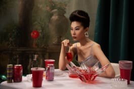 Une femme asiatique avec une coiffure élégante et des boucles d'oreilles originales, assise à une table avec des verres et des canettes, tenant un verre de liquide rouge, avec un fond de jardin peint et un petit robot 