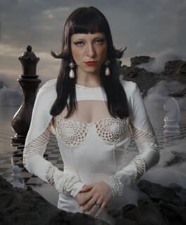 Portrait d'une femme aux longs cheveux noirs et robe blanche élégante, posant devant un fond de plateau d'échecs en 3D avec des pièces géantes et des nuages. 